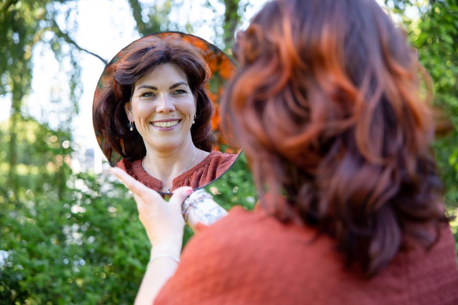 Petra Scholten kijkt glimlachend naar haar spiegelbeeld in een handspiegel, buiten tussen het groen